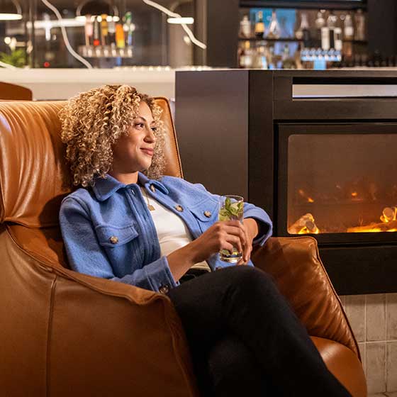 Une invitée tenant un verre et assise en face du bar dans un fauteuil du salon d’aéroport