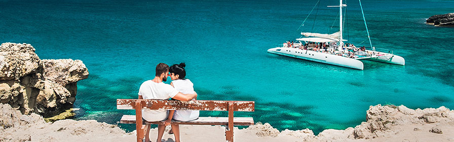 Couple on a bench in a tropical location. Catamaran in the water in the background.