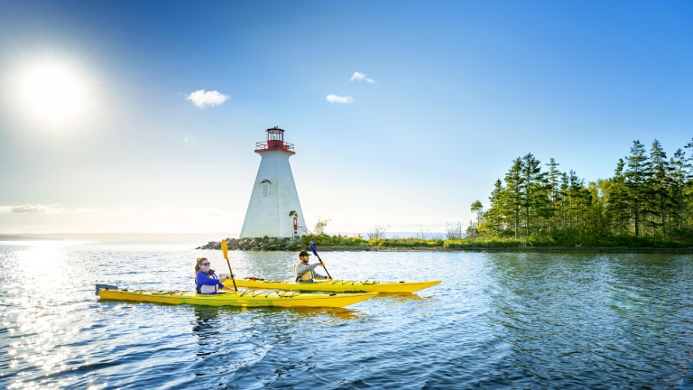 Two kayakers paddle past lighthouse