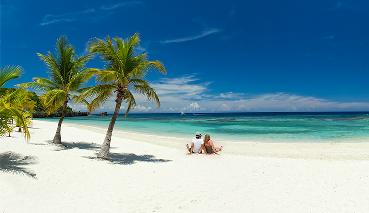 Man and woman looking at ocean while sitting on white sand beach