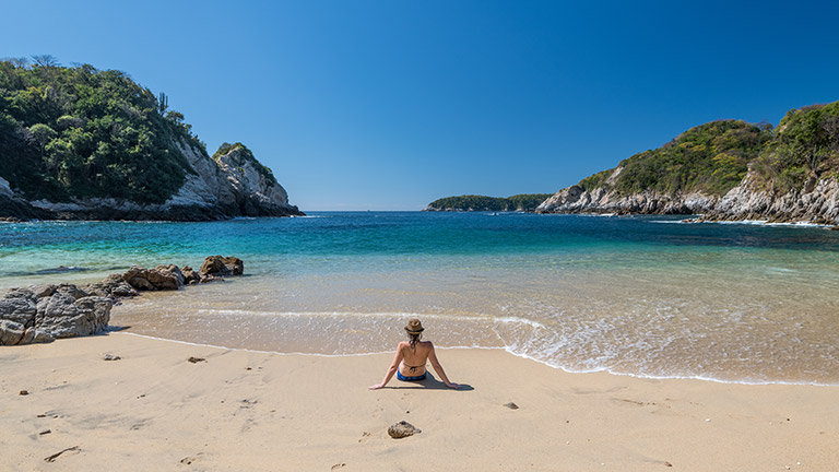 A woman sitting on a sandy beach looking out towards the sea in Huatulco, Mexico.